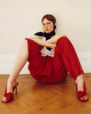 Model seated on parquet floor wearing red trousers, white top, and red heeled sandals with black neck scarf