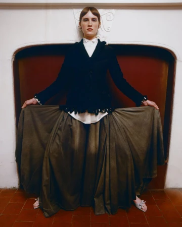 Model holding out a wide olive-green maxi skirt while posing in front of an arched white fireplace alcove.