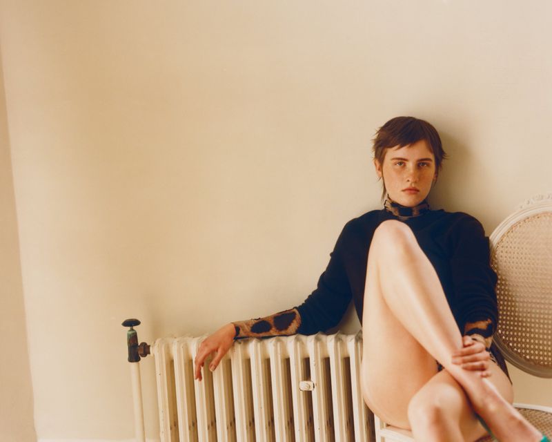 Woman with short brown hair seated on a vintage cane chair beside a cast iron radiator against a cream wall.