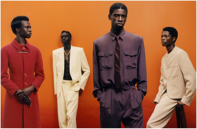 Four male models posing against a solid orange wall wearing tailored garments in red, white, purple, and cream.