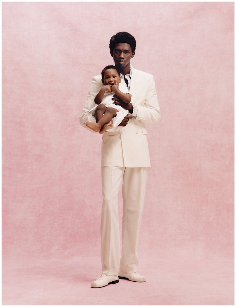 A Black man in a cream tailored suit holding a toddler in white against a textured blush pink studio background.