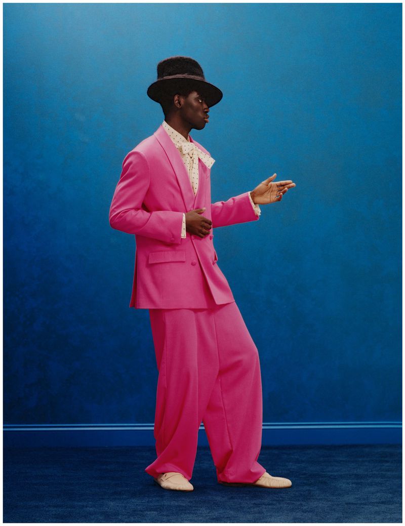 Black male model wearing an oversized bright pink suit, dark fedora, and patterned neck bow against a deep blue wall.