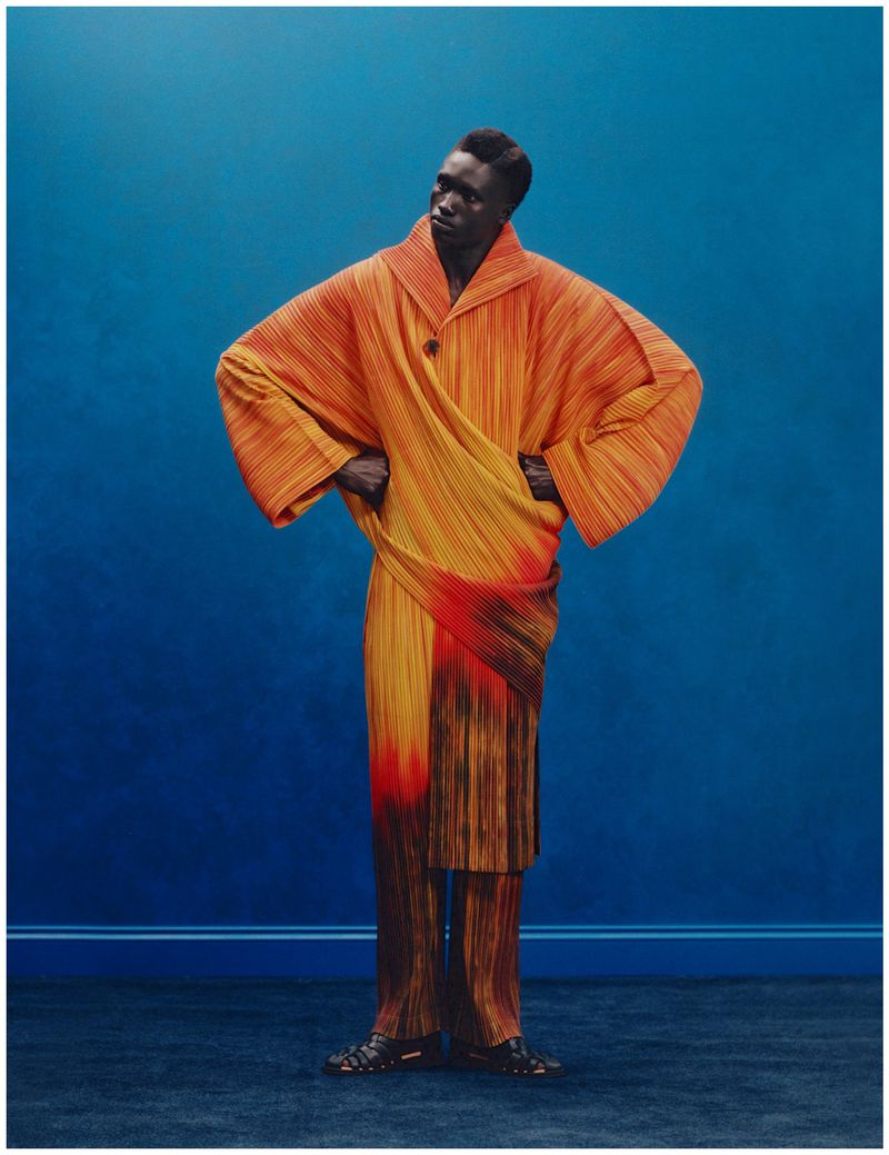 Black man wearing a pleated orange and yellow robe standing against a solid blue studio backdrop for Glass Man Dandy Diaspora