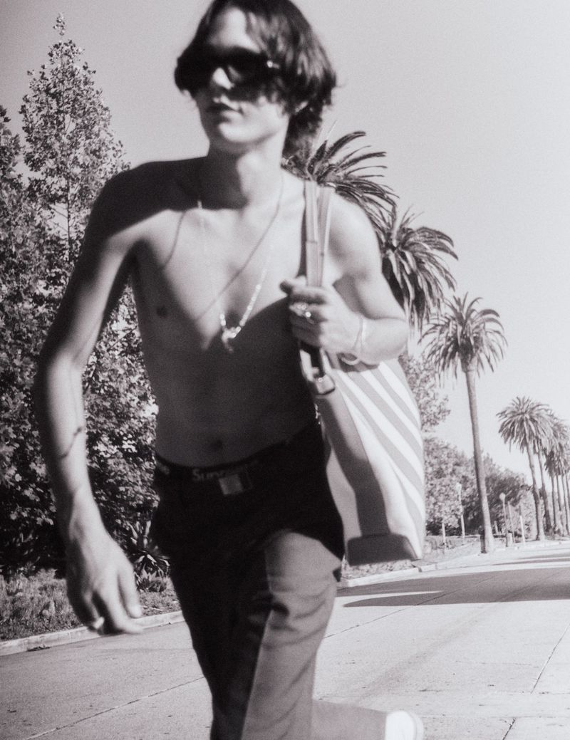 Shirtless man in sunglasses and dark trousers walking down a palm-lined street carrying a striped tote bag.