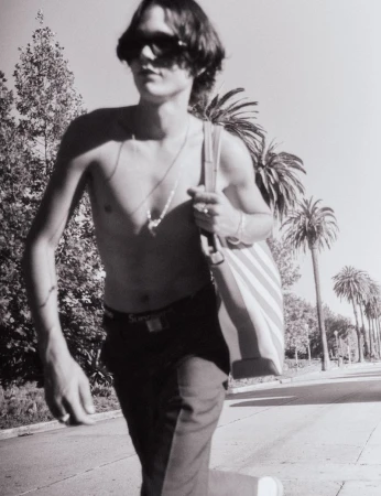 Shirtless man in sunglasses and dark trousers walking down a palm-lined street carrying a striped tote bag.