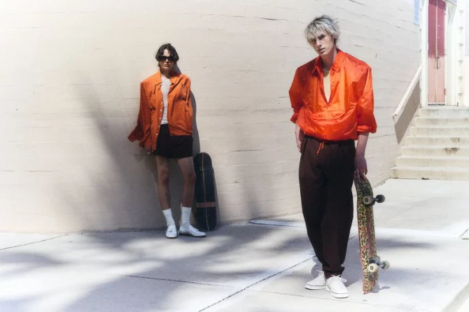 Two male models posing with skateboards in bright sunlight against a curved concrete wall, wearing vibrant orange streetwear tops.