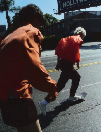 Two young men skateboard down a sunlit paved road under a Hollywood billboard, wearing orange and terracotta jackets.