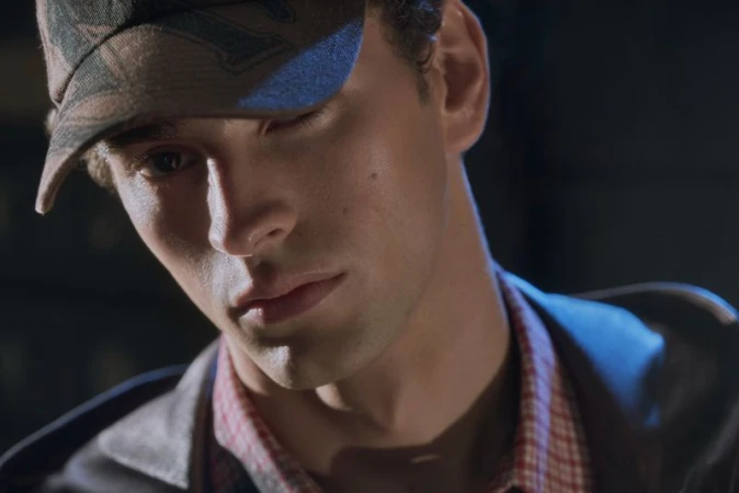 Close-up portrait of a young man looking down while wearing a camouflage baseball cap and a red patterned collared shirt.