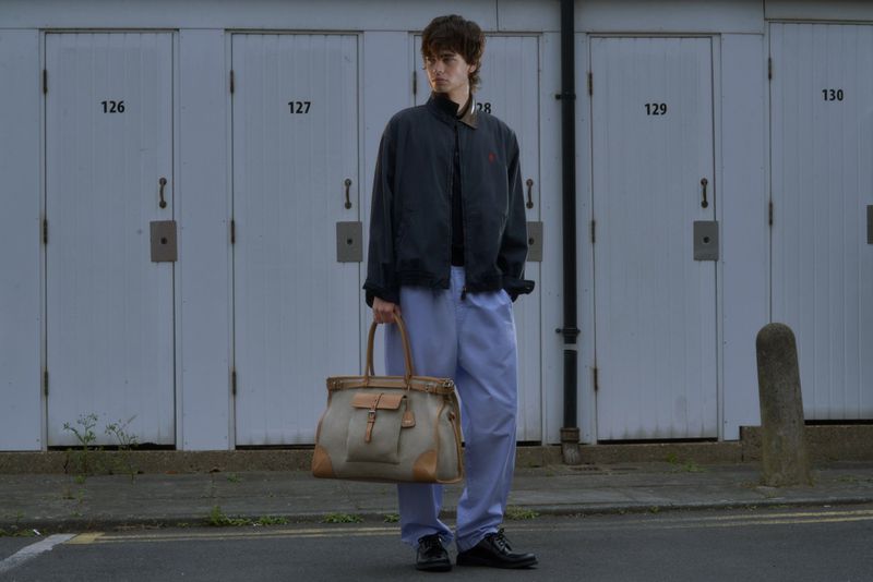 Male model holding a large beige canvas weekend bag in front of a row of numbered white garage doors.