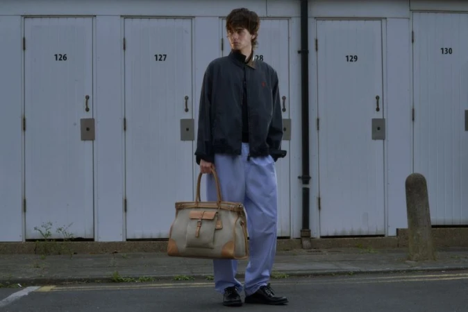 Male model holding a large beige canvas weekend bag in front of a row of numbered white garage doors.