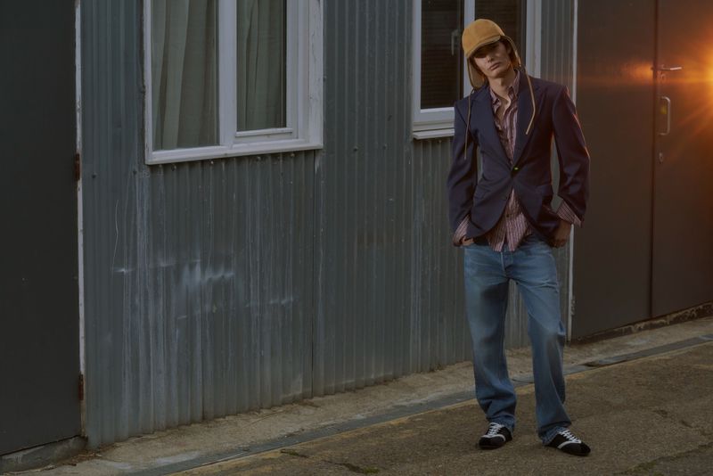 Male model in a navy blazer, yellow flap hat, and blue denim jeans standing outdoors beside a corrugated metal building.