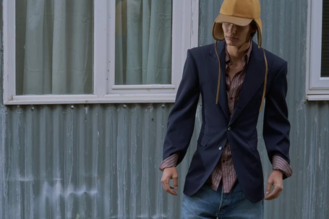 Male model wearing a navy blazer, striped shirt, blue jeans, and a tan trapper hat against a corrugated metal wall.