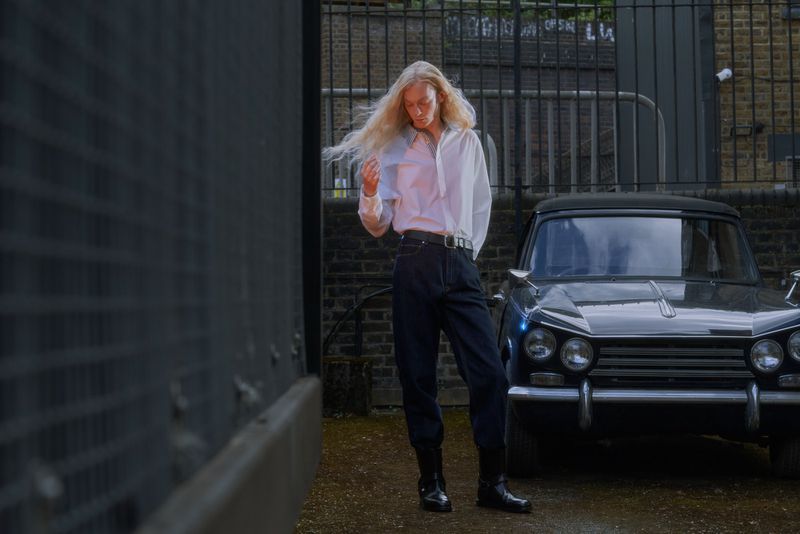 Model with long blonde hair in a white shirt and dark trousers standing beside a parked vintage car outdoors.