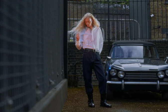 Model with long blonde hair in a white shirt and dark trousers standing beside a parked vintage car outdoors.