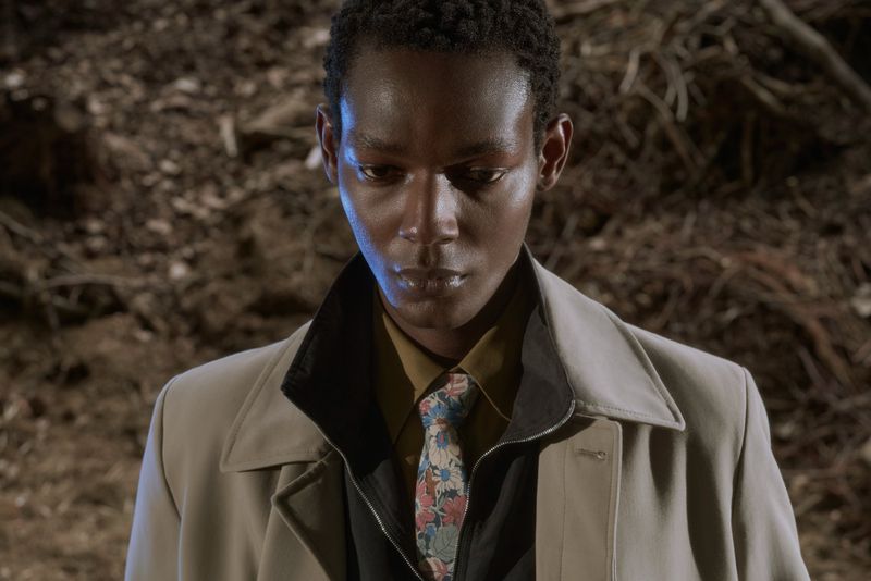 Close-up portrait of a Black man wearing a beige overcoat, dark jacket, and floral patterned tie against a woodsy ground background.