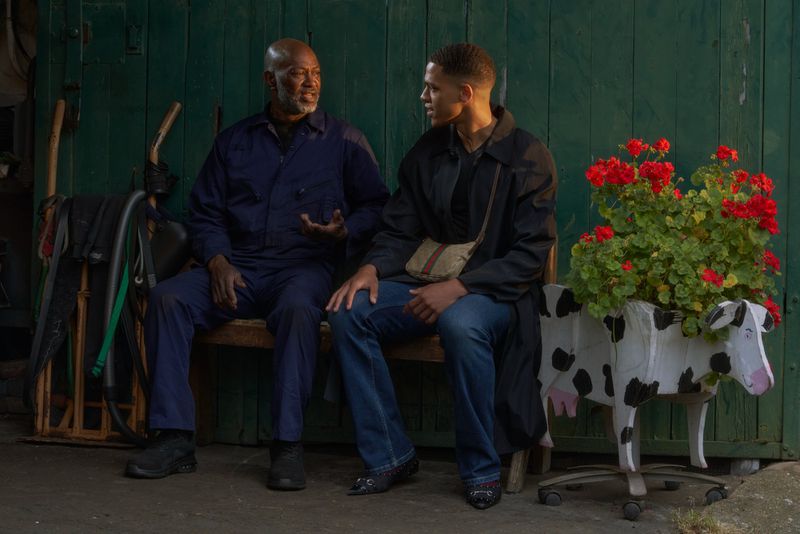 Two men seated on a wooden bench talking in front of a green wooden door beside a cow shaped flower planter.
