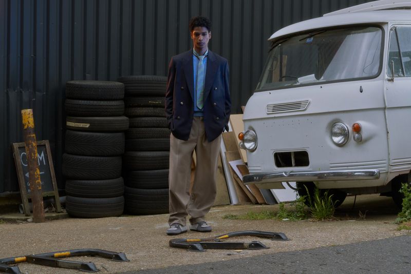 Model wearing a navy double-breasted blazer and wide-leg beige trousers standing outdoors beside a vintage white van and stacked tires.
