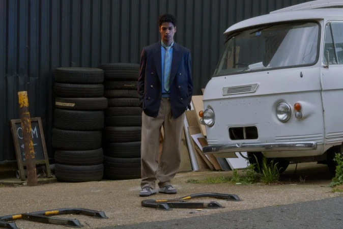 Model wearing a navy double-breasted blazer and wide-leg beige trousers standing outdoors beside a vintage white van and stacked tires.