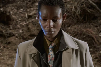 Close-up portrait of a Black man wearing a beige overcoat, dark jacket, and floral patterned tie against a woodsy ground background.