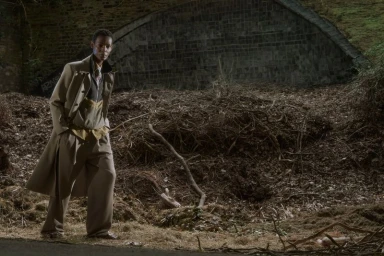 Model wearing a tailored beige trench coat and wide-leg trousers outdoors in front of a stone arch and brush landscape.