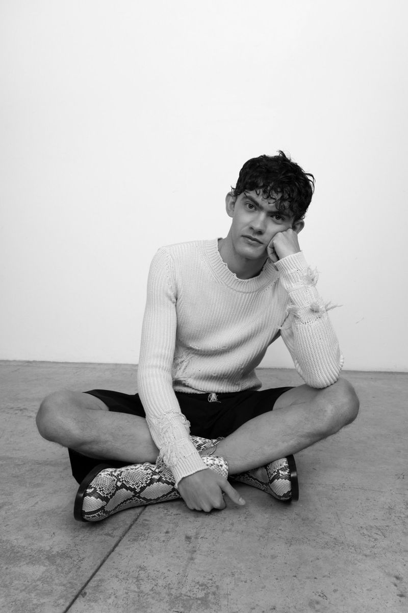 Portrait of Joe Locke sitting crossed-legged on the floor wearing a light sweater and patterned boots against a white wall.
