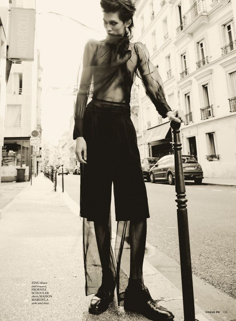 Woman in sheer top and culottes stands on a city street, leaning on a pole, with neutral color palette and magazine editorial style.