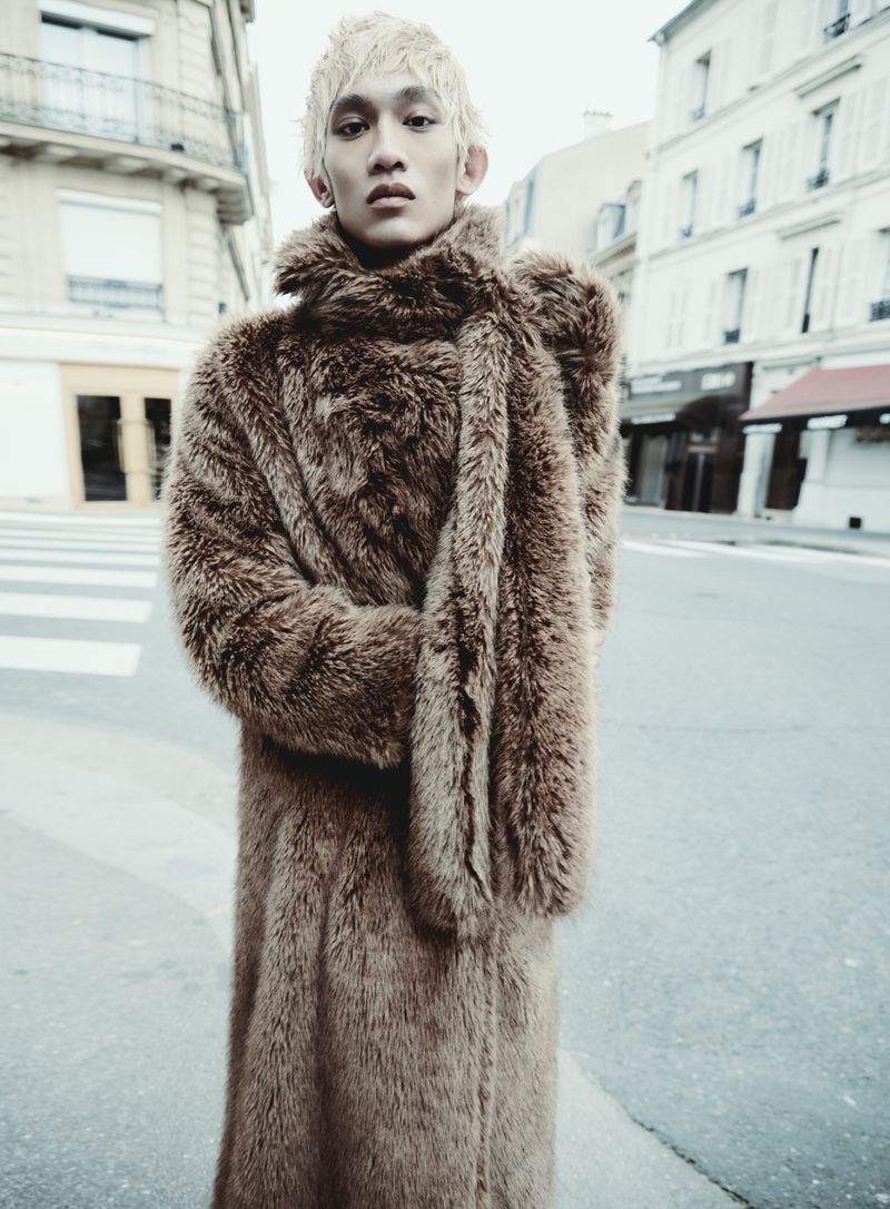 Asian male model in long brown fur coat on urban street with apartment buildings in the background; fashion editorial shot in outdoor setting.