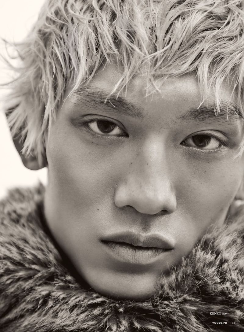 Close-up portrait of an Asian male model with fair hair and a fur collar, gazing intently, in a studio environment.