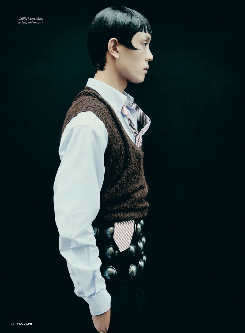 Side-view portrait of a young adult male with unique hairstyle, wearing a collared shirt, vest, and studded belt against a dark backdrop.