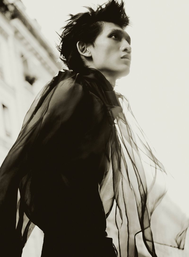 Monochrome portrait of a slender male model with spiky hair, wearing a sheer ruffled top against an outdoor urban building backdrop, gazing upward.
