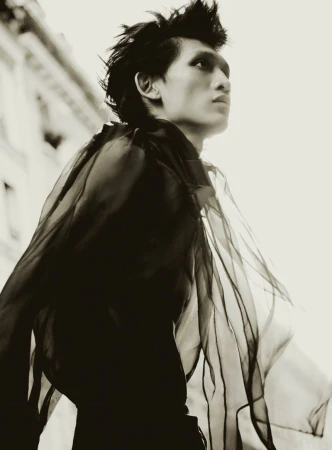 Monochrome portrait of a slender male model with spiky hair, wearing a sheer ruffled top against an outdoor urban building backdrop, gazing upward.