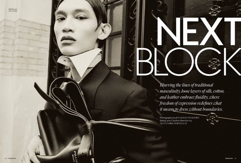 Fashion editorial featuring a stylishly dressed adult male with Prada bag, posing near wrought iron gate against an off-white brick building.