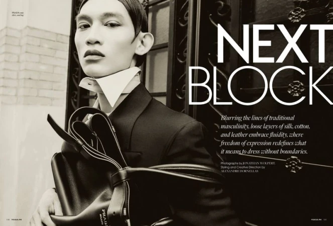 Fashion editorial featuring a stylishly dressed adult male with Prada bag, posing near wrought iron gate against an off-white brick building.