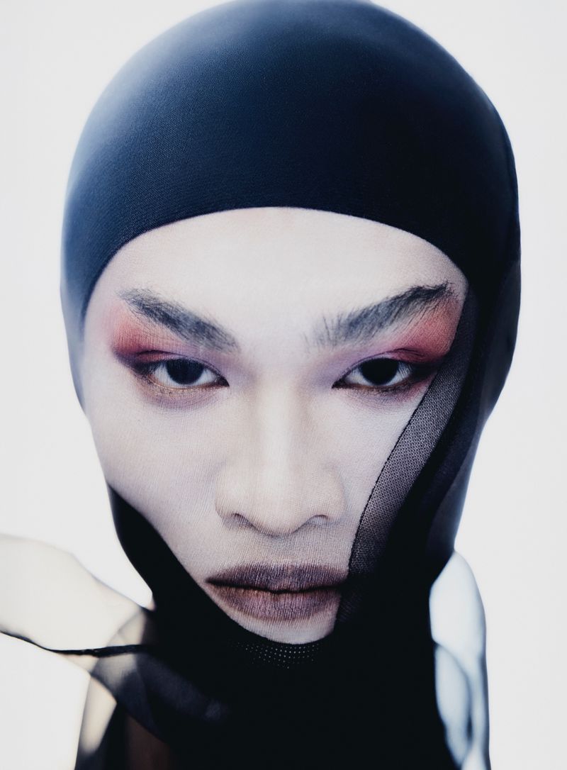 Close-up portrait of a person with artistic makeup and head covering against a white background, emphasizing bold makeup and unique style.