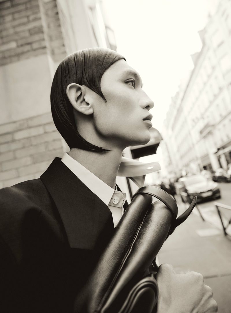 Monochrome portrait of an adult with short dark hair, wearing a dark suit with a white shirt, and holding a dark bag in a city street.