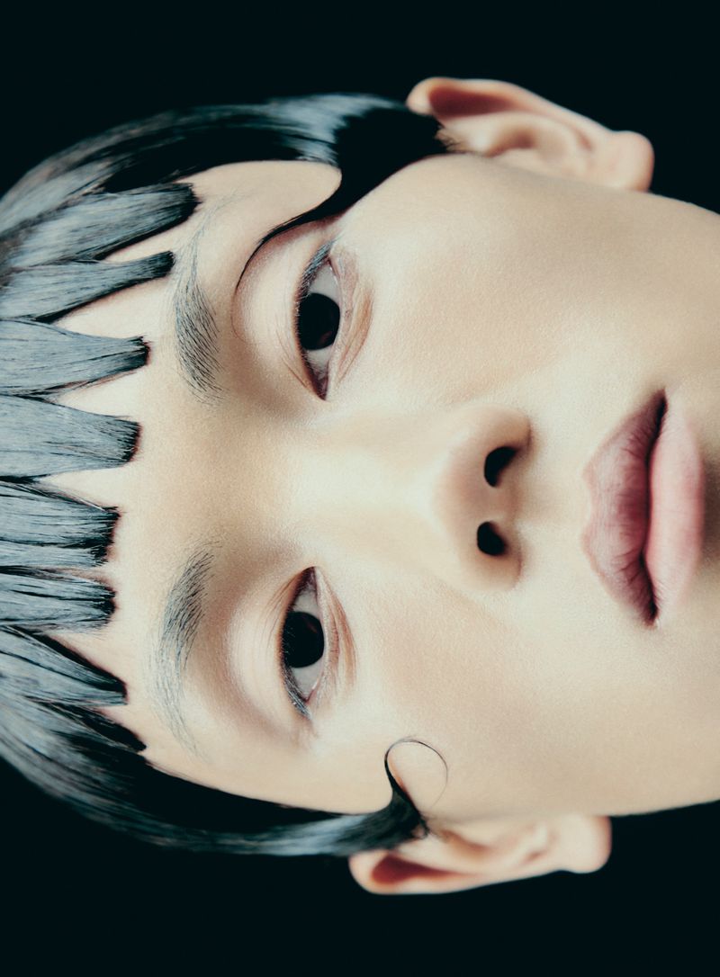 Close-up portrait of a person with black hair styled in zig-zag patterns across their forehead against a dark background.
