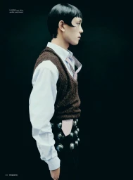 Model in profile wearing a white button-down shirt, brown knit vest, and studded black trousers against a dark backdrop.