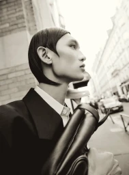 Eden Huang photographed in profile wearing a black tailored coat, white shirt, and carrying a black leather bag on a city street.