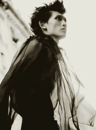 Model Eden Huang in a sheer black flowing garment looking upwards against an outdoor architectural background.