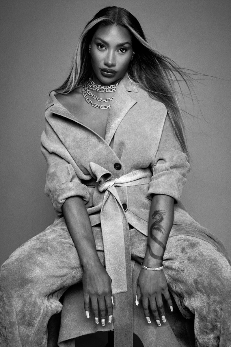 Aya Nakamura seated wearing a belted coat, layered chain necklace, and showing a forearm tattoo in a black-and-white studio portrait.