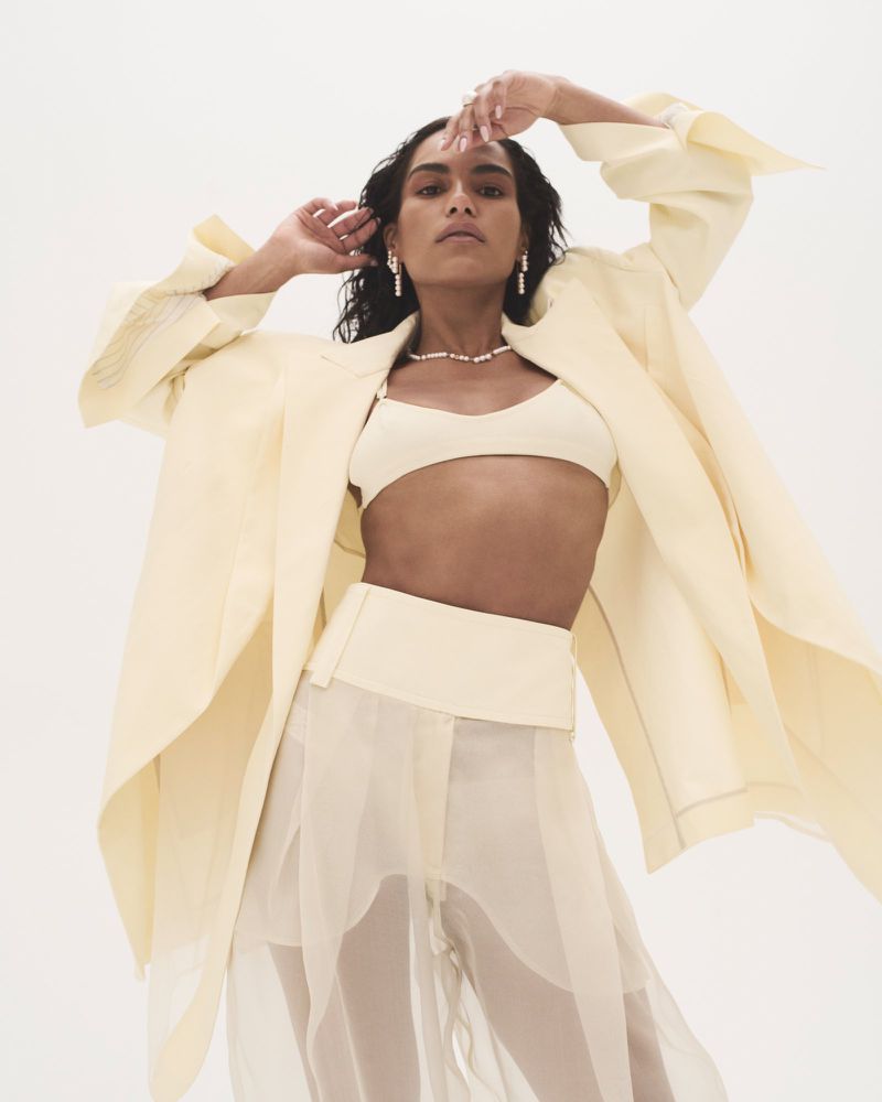 Saffron Hocking posing in an oversized pale yellow coat, matching bralette, and sheer trousers against a white seamless background.