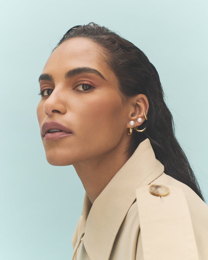 Saffron Hocking in a beige trench coat wearing pearl and gold earrings against a light blue studio backdrop.