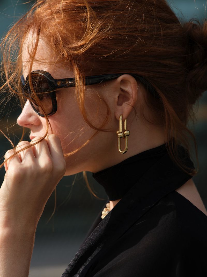 Profile portrait of Ellie Bamber wearing dark sunglasses, gold link earrings, and a black high-neck top with red hair in an updo.