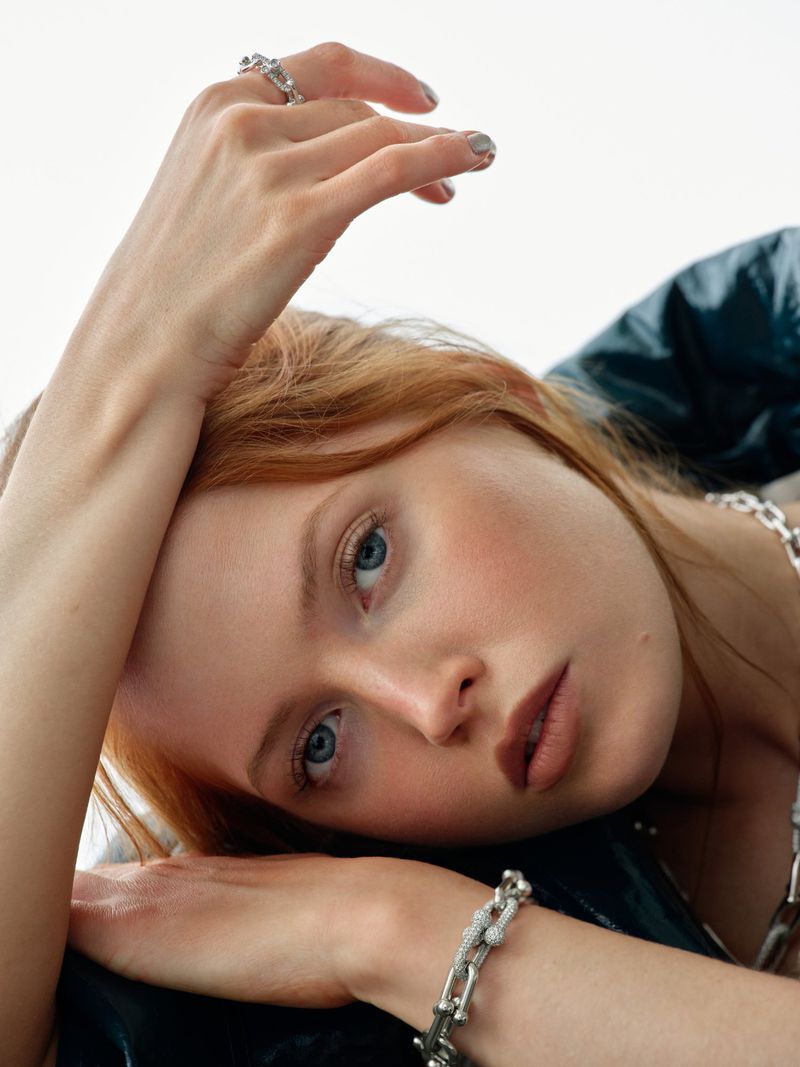 Close-up portrait of actress Ellie Bamber with red hair resting her hand near her face wearing silver jewellery against a white backdrop.