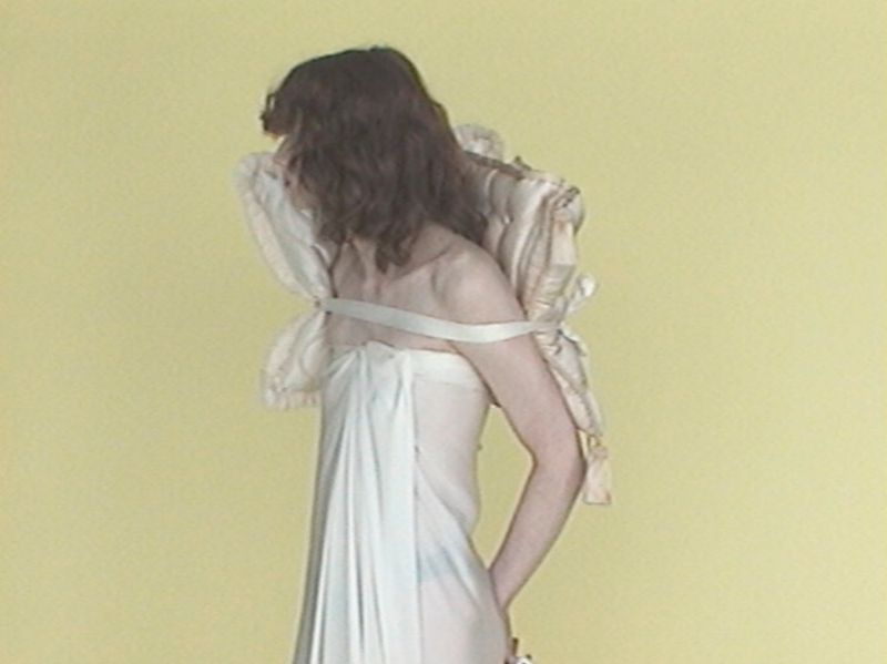 Model with wavy brown hair wearing a draped white fabric dress and a strapped cream quilted cushion on their back against a yellow wall.