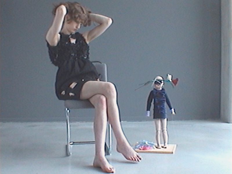 Model seated on a chair in a black dress next to a small doll figure against a grey studio backdrop.