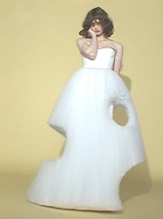 Model poses in a sculptural white tulle ballgown with abstract cutout shapes against a pale yellow studio background.