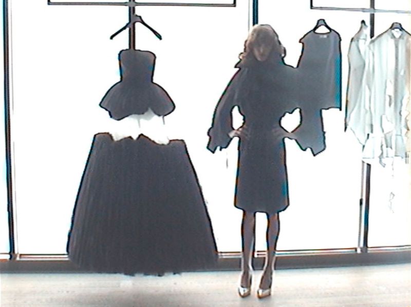 Model standing with hands on hips beside a large hanging black strapless gown in front of bright windows.