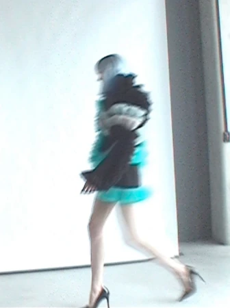 Motion-blurred model walking in black high heels, a dark ruffled jacket, and bright teal garment against a white studio wall.