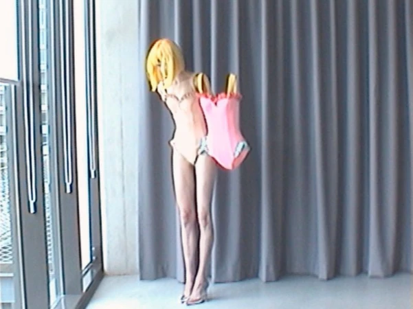 Model wearing a blonde wig and nude bodysuit posing with an attached pink swimwear cutout against grey curtains.